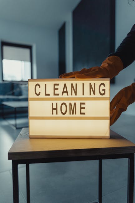 A person wearing orange rubber cleaning gloves is handling an illuminated lightbox sign displaying the words 'CLEANING HOME' on a wooden table in a modern, well-lit living room. The background feature